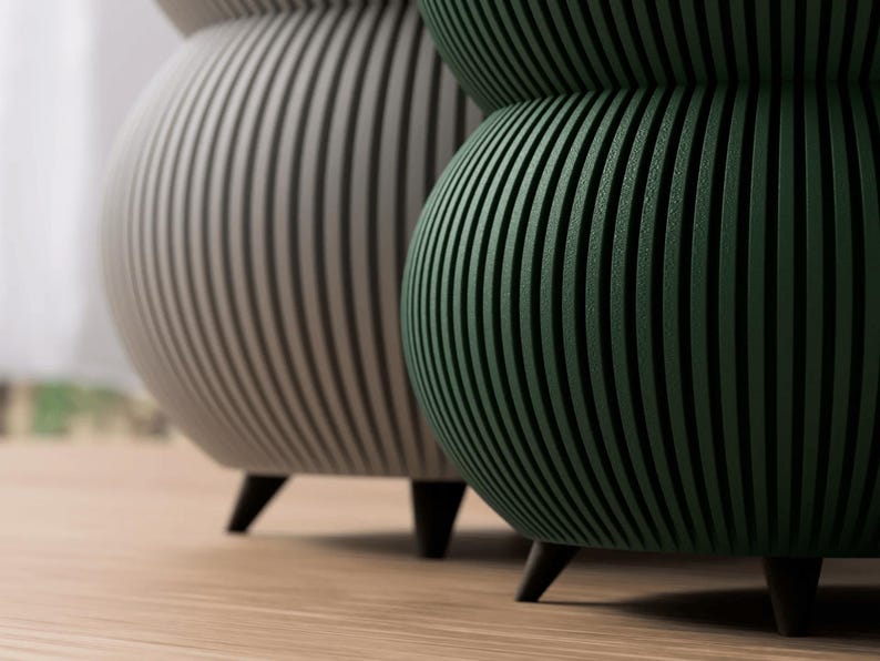 May include: Two decorative, modern accent tables. One is a light gray color, and the other is a dark green. Both have a ribbed design and sit on three black legs. The tables are on a light brown wooden floor.