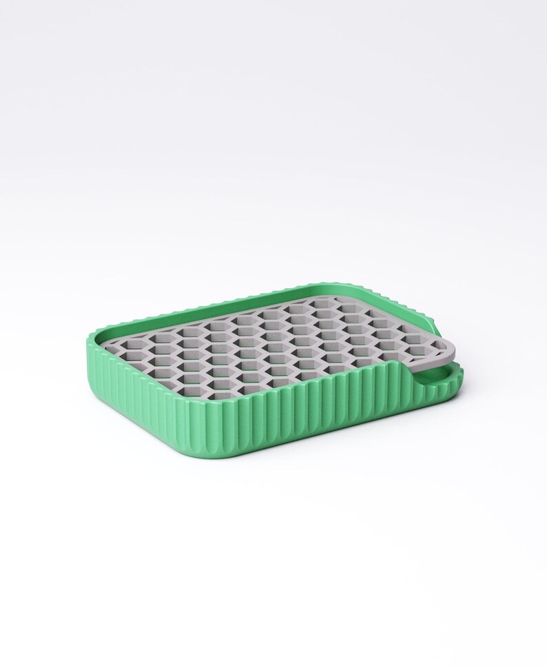 May include: A green and gray silicone ice cube tray. The tray has a rectangular shape with a ribbed green border and a gray honeycomb-patterned interior. A small tab extends from one side.