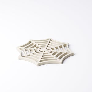May include: A light beige, spider web-shaped trivet. The trivet is made of a solid material and features a detailed web design. The trivet is designed to protect surfaces from hot items. A decorative item for the kitchen.