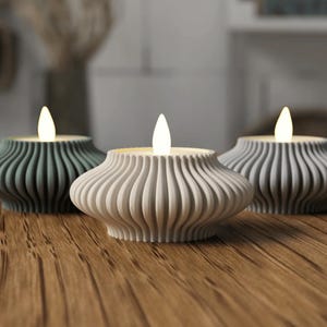 Puede incluir: Tres velas LED decorativas con un dise&ntilde;o ondulado y acanalado. Las velas son en tonos grises y blancos, con una luz c&aacute;lida y parpadeante. Est&aacute;n dispuestas sobre una superficie de madera, creando un ambiente acogedor.