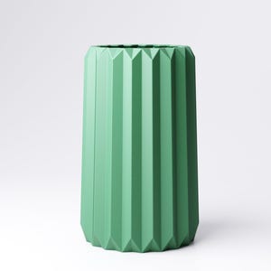 May include: A tall, cylindrical vase in a muted green hue. The vase features a geometric design with vertical, ridged facets that run the length of the body. The vase has a modern, minimalist aesthetic, suitable for home decor.