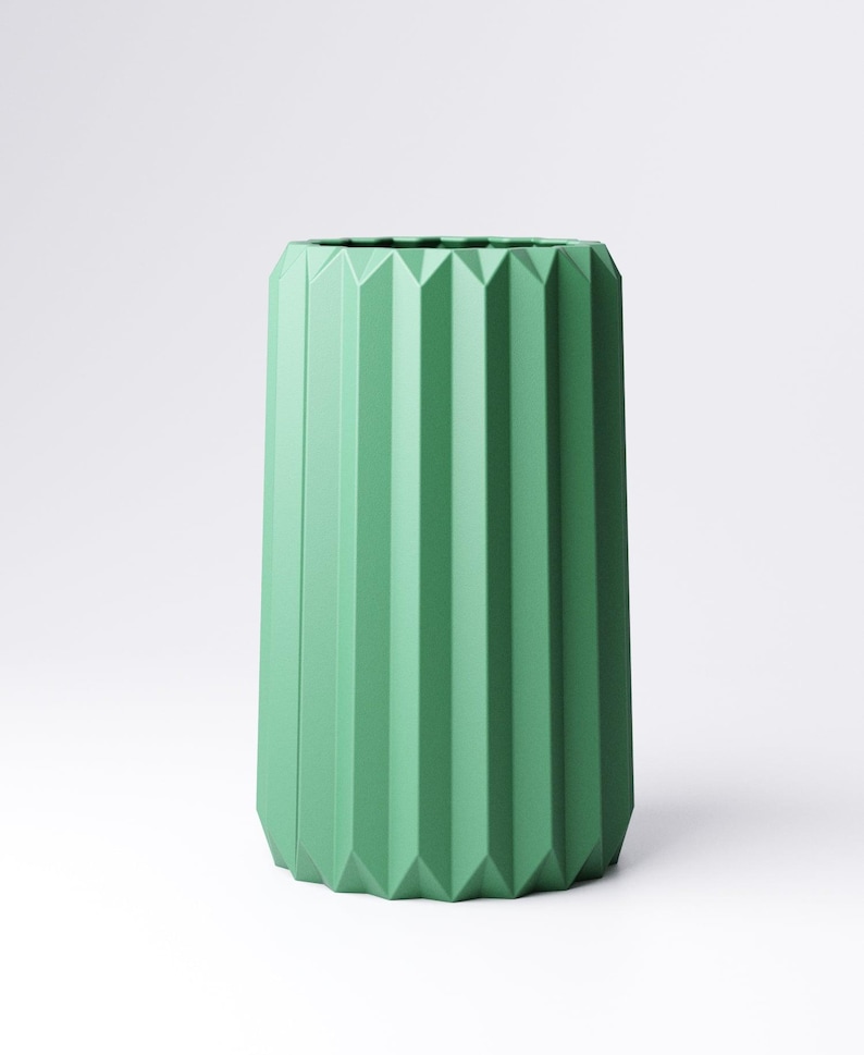 May include: A tall, cylindrical vase in a muted green hue. The vase features a geometric design with vertical, ridged facets that run the length of the body. The vase has a modern, minimalist aesthetic, suitable for home decor.