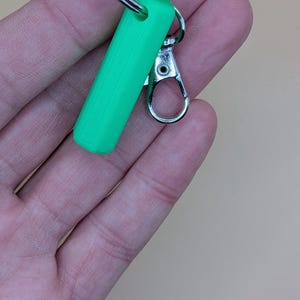 May include: A bright green rectangular keychain fob with a silver-colored clasp and key ring. The fob is a solid, smooth shape. The clasp is open, ready to attach to a bag or other item.