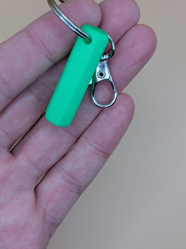 May include: A bright green rectangular keychain fob with a silver-colored clasp and key ring. The fob is a solid, smooth shape. The clasp is open, ready to attach to a bag or other item.
