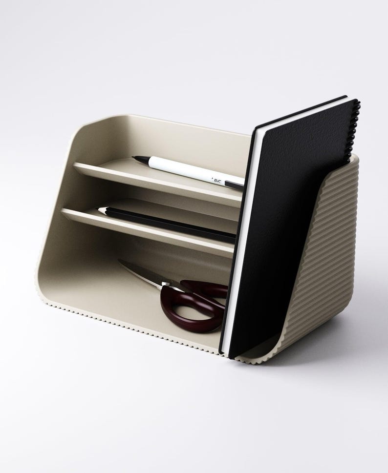 May include: A beige desk organizer with two shelves, holding a black notebook, a white pen, a black pencil, and burgundy scissors. The organizer has a ribbed side and a dotted edge, providing a stylish storage solution for office supplies.