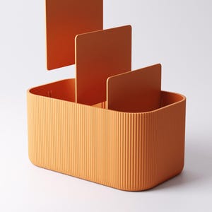 May include: An orange desk tidy with a rectangular base and vertical ridged texture. Three orange dividers are placed inside the tidy. The tidy is designed to hold office supplies.
