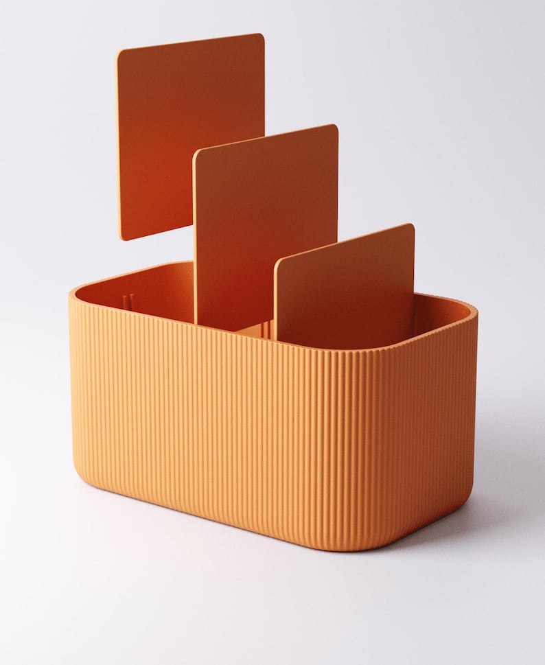 May include: An orange desk tidy with a rectangular base and vertical ridged texture. Three orange dividers are placed inside the tidy. The tidy is designed to hold office supplies.