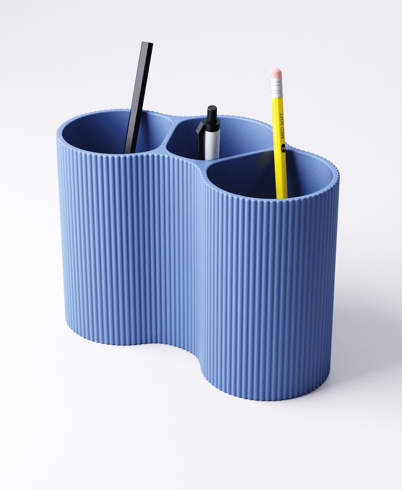 May include: A blue desk organizer with a wavy design, holding a black pen, a silver and black pen, and a yellow pencil. The organizer has a ribbed texture and is made of a solid, opaque material. The pencil has a pink eraser.