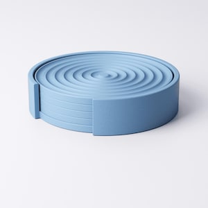 May include: A set of six light blue coasters, each with a ripple pattern on the top. The coasters are arranged in a circular stack, with a rectangular side piece for easy removal. The coasters are made of a solid material.