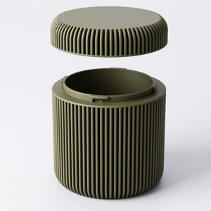 May include: Olive green cylindrical container with a matching lid. The container and lid have vertical ribbed detailing. The lid is slightly elevated above the container, suggesting it is removable. The background is plain white.