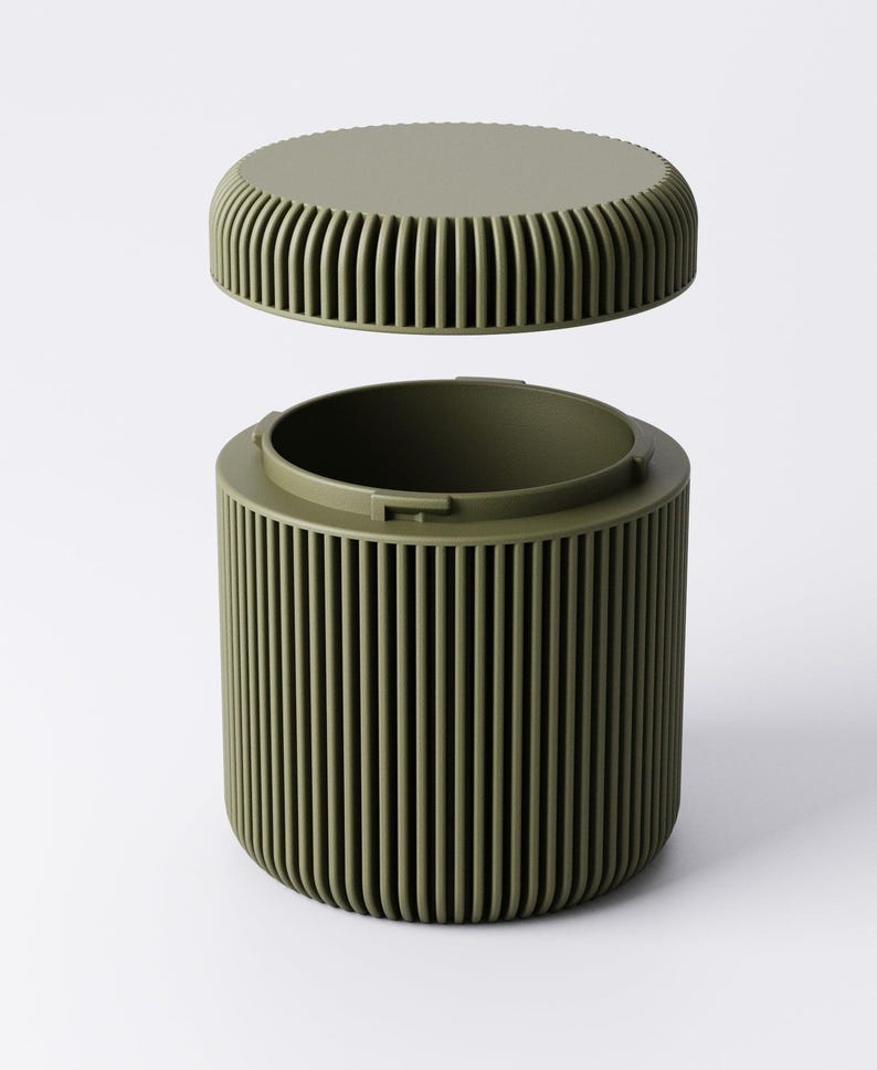 May include: Olive green cylindrical container with a matching lid. The container and lid have vertical ribbed detailing. The lid is slightly elevated above the container, suggesting it is removable. The background is plain white.