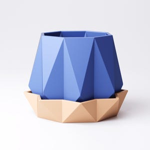 May include: A geometric blue planter with a matching beige saucer. The planter has a faceted design and a hexagonal top. The saucer is also faceted, creating a modern, minimalist aesthetic. The planter and saucer are made of a solid material.