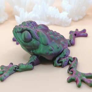 May include: A 3D-printed frog figurine in shades of green and purple. The frog has a textured surface and articulated limbs, with a detailed design. The frog is posed on a light-colored surface, with white, crystalline shapes in the background.