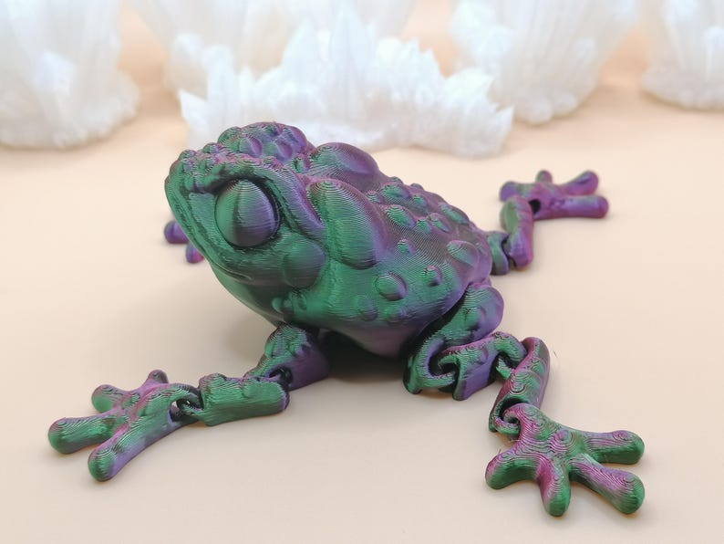 May include: A 3D-printed frog figurine in shades of green and purple. The frog has a textured surface and articulated limbs, with a detailed design. The frog is posed on a light-colored surface, with white, crystalline shapes in the background.