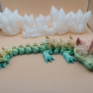 Puede incluir: Un juguete de drag&oacute;n articulado multicolor, con un degradado de colores que va del verde al azul y al rosa. El drag&oacute;n est&aacute; sobre una superficie beige, con formaciones de cristales blancos en el fondo.