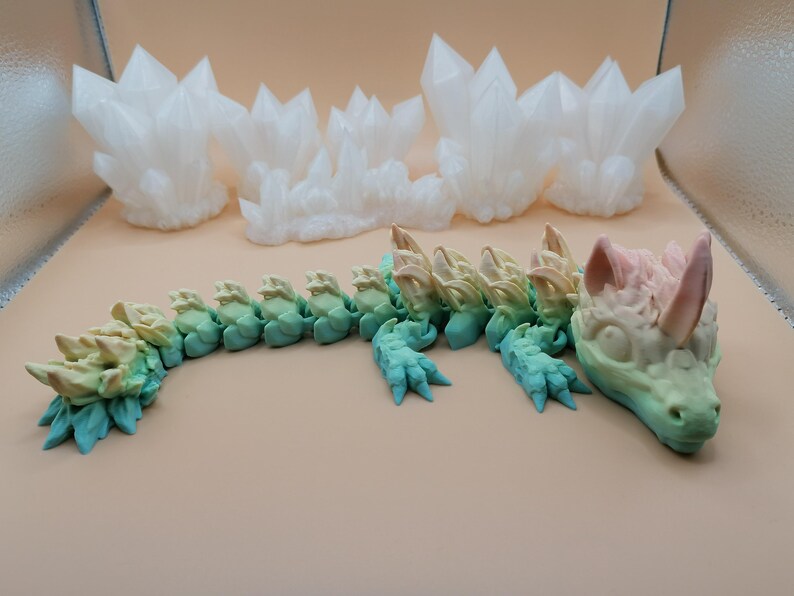 Puede incluir: Un juguete de drag&oacute;n articulado multicolor, con un degradado de colores que va del verde al azul y al rosa. El drag&oacute;n est&aacute; sobre una superficie beige, con formaciones de cristales blancos en el fondo.