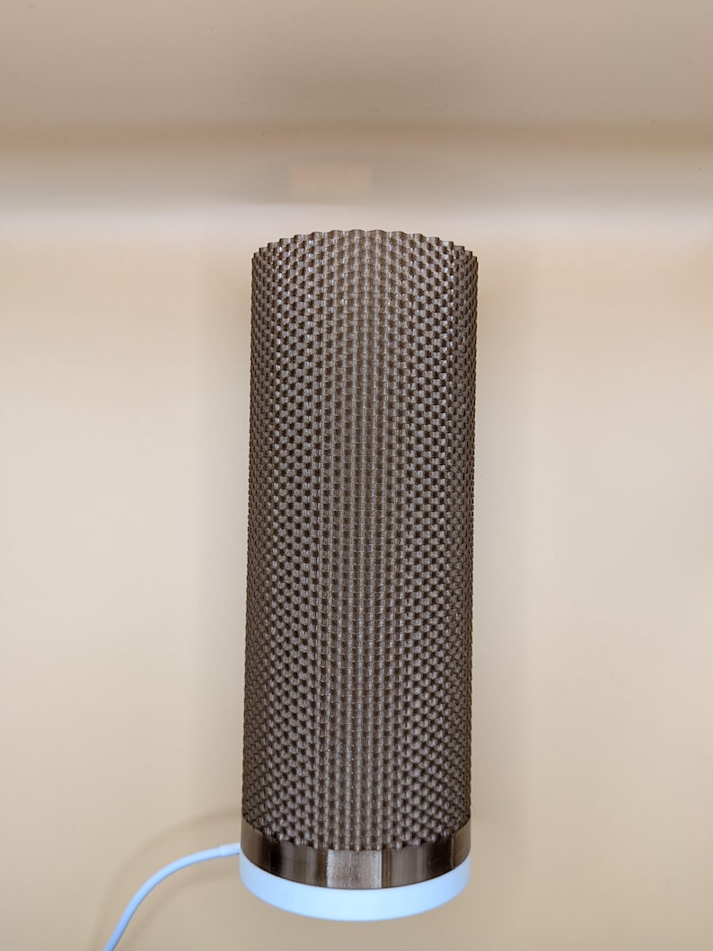 K&ouml;nnte beinhalten: Eine zylindrische Lampe mit einer strukturierten, dunkelbraunen Au&szlig;enseite. Die Lampe hat ein silbernes Band am Sockel, &uuml;ber einem hellblauen Abschnitt. Ein blaues Kabel erstreckt sich von der Basis. Die Lampe steht vor einer beigen Wand.