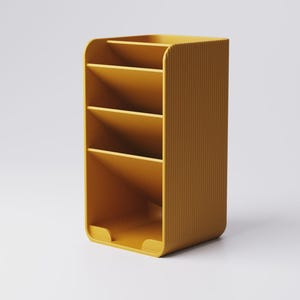May include: Mustard yellow desk organizer with four angled compartments and a curved design. The organizer has a ribbed texture on the side and a small lip at the base. The product is designed to hold office supplies.