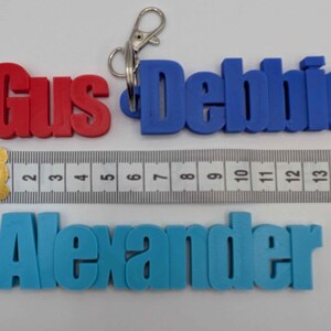 May include: Three personalized keychains in red, blue, and light blue, spelling out "Gus", "Debbie", and "Alexander". A ruler shows measurements in inches. Each keychain has a metal clasp for attaching to keys or bags.