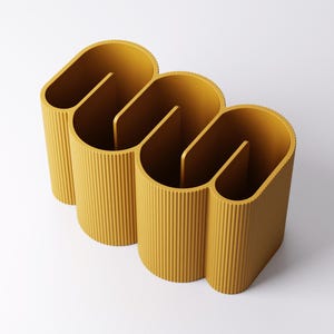 May include: Mustard yellow desk organizer with a unique, wavy design. The organizer has three sections, each with a vertical divider. The exterior features vertical ribbed detailing. The product is made of a solid material and sits on a white surface.
