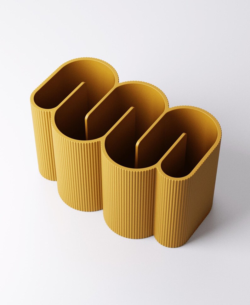 May include: Mustard yellow desk organizer with a unique, wavy design. The organizer has three sections, each with a vertical divider. The exterior features vertical ribbed detailing. The product is made of a solid material and sits on a white surface.