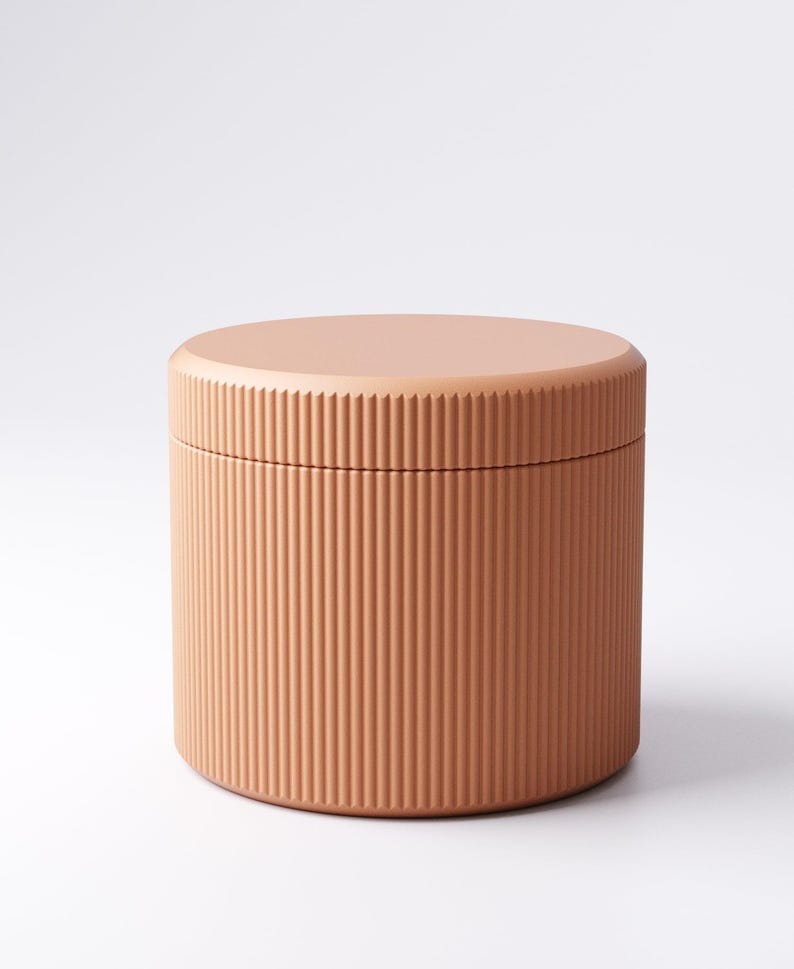 May include: A cylindrical storage container with a lid. The container is a solid peach color with vertical ribbed detailing. The lid is flat and round, matching the container's color. The container is likely made of ceramic or a similar material.