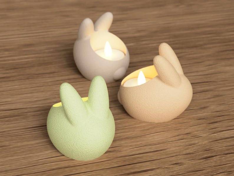 K&ouml;nnte beinhalten: Drei hasenf&ouml;rmige Kerzenhalter in Wei&szlig;, Hellgr&uuml;n und Beige, jeweils mit einer brennenden Kerze. Die Halter stehen auf einer Holzoberfl&auml;che und erzeugen eine warme Atmosph&auml;re. Ideal f&uuml;r Ostern oder als Dekoration.