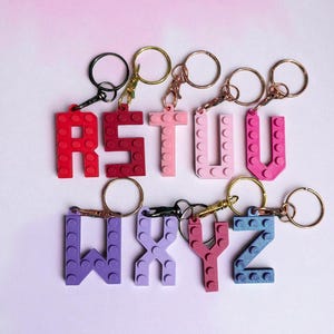 May include: A collection of colorful letter keychains, each crafted from interlocking brick-style pieces. The letters R, S, T, U, W, X, Y, and Z are displayed in shades of red, pink, purple, and blue, with gold and black key rings.
