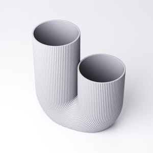 K&ouml;nnte beinhalten: Eine hellgraue U-f&ouml;rmige Vase mit einer vertikal gerippten Textur auf der Au&szlig;enseite. Das Objekt besteht aus zwei zylindrischen &Ouml;ffnungen unterschiedlicher H&ouml;he, die durch einen geschwungenen Boden verbunden sind und eine geriffelte Oberfl&auml;che bilden.