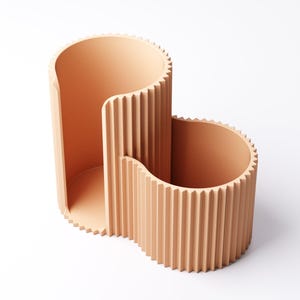 May include: A peach-colored, modern desk organizer with a unique, wavy design. The organizer features two open compartments of varying heights, each with vertical ridged detailing. The top edges have a sawtooth pattern. The minimalist design is suitable for office or home use.