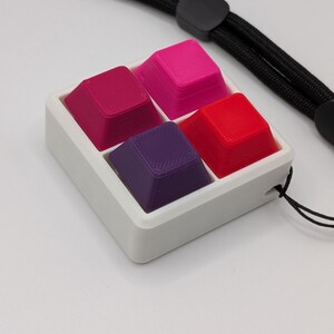 May include: A white square container holds four colorful keycaps: magenta, hot pink, red, and purple. A black lanyard is attached to the container. The keycaps are arranged in a 2x2 grid.
