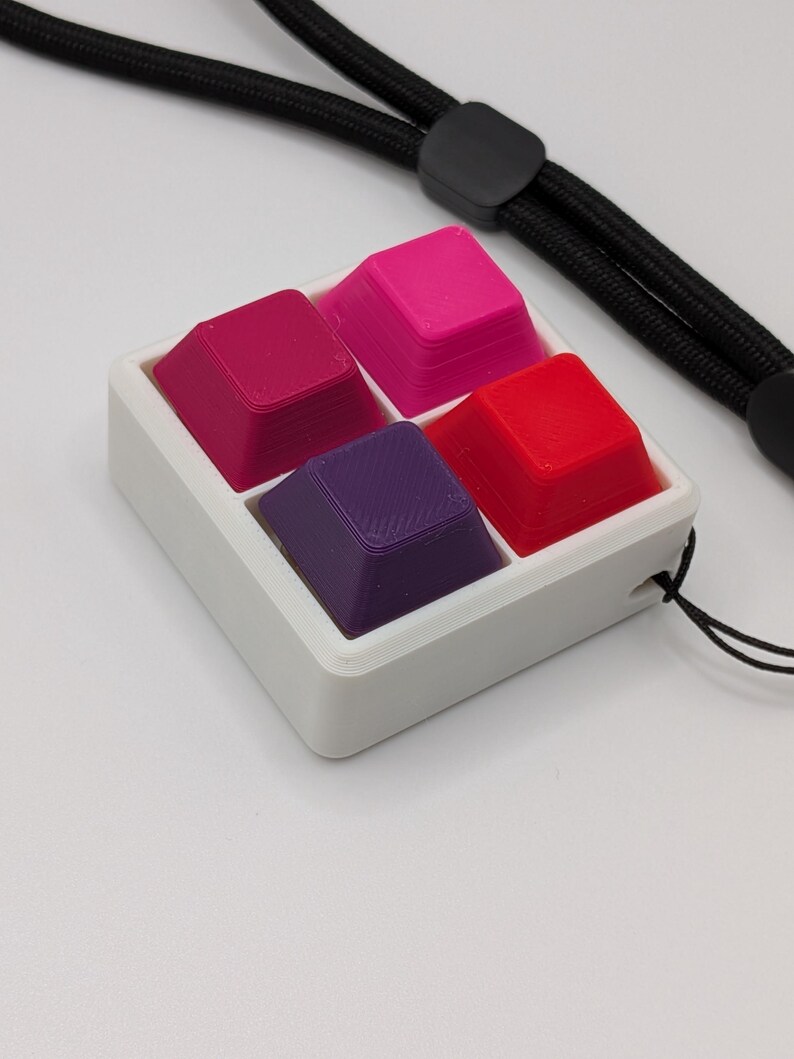 May include: A white square container holds four colorful keycaps: magenta, hot pink, red, and purple. A black lanyard is attached to the container. The keycaps are arranged in a 2x2 grid.