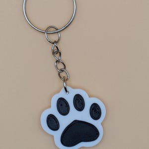 May include: A white and black paw print keychain with a silver keyring and chain. The paw print design features a white background with black details, and is likely made of plastic or a similar material.