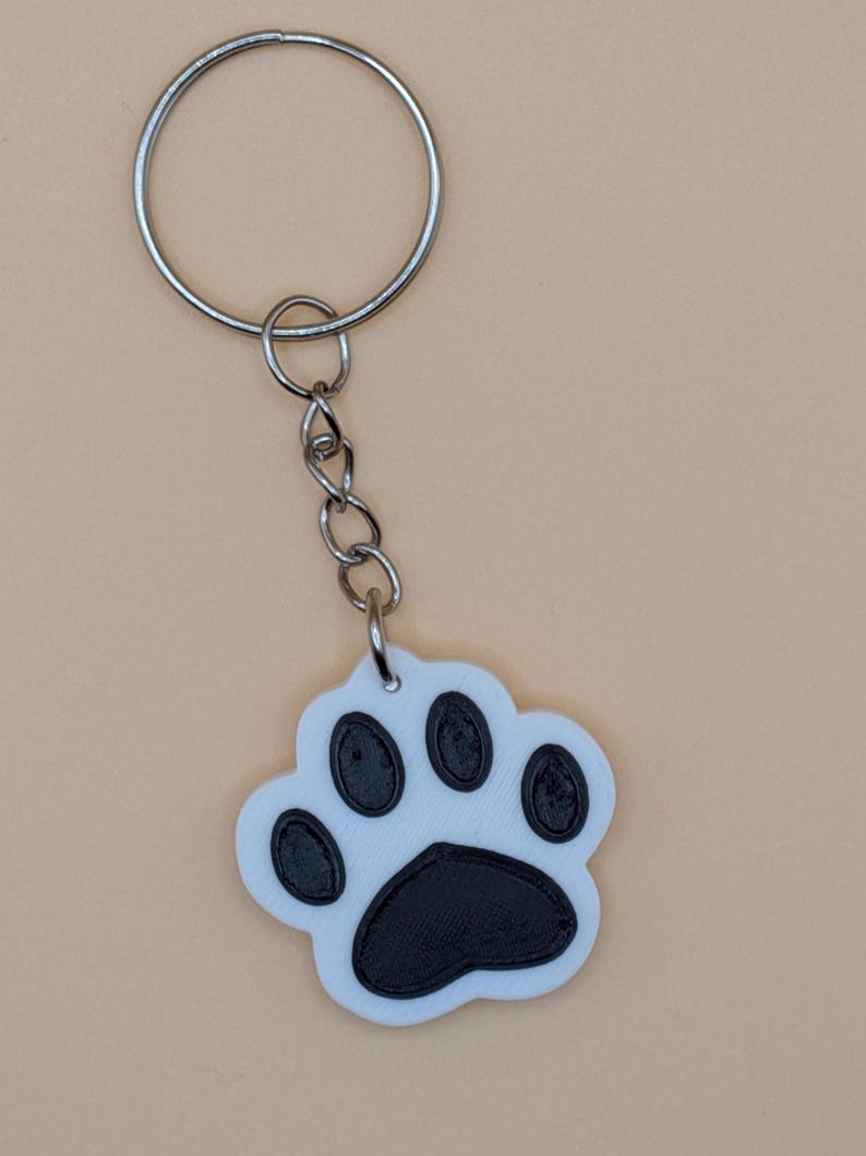 May include: A white and black paw print keychain with a silver keyring and chain. The paw print design features a white background with black details, and is likely made of plastic or a similar material.