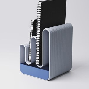 May include: A modern desk organizer in shades of gray and blue, holding three spiral-bound notebooks. The organizer has a curved, ribbed design and a blue base, providing a stylish storage solution for office or home use.