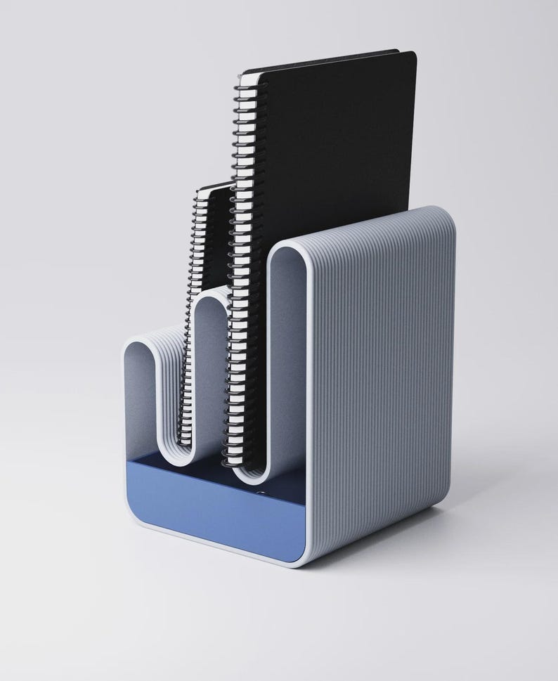 May include: A modern desk organizer in shades of gray and blue, holding three spiral-bound notebooks. The organizer has a curved, ribbed design and a blue base, providing a stylish storage solution for office or home use.