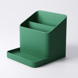 May include: A solid green desk organizer with a ribbed texture. The organizer has a rectangular base with a small tray, a main compartment with a divider, and a slanted back. The overall design is minimalist and modern.