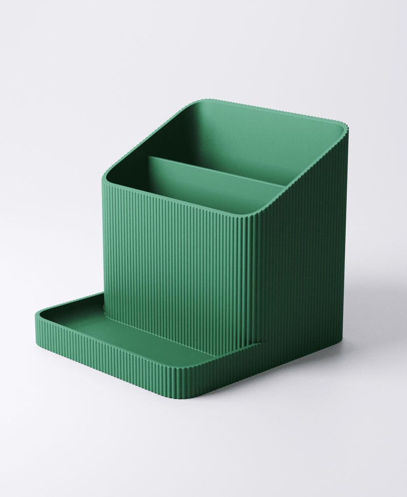 May include: A solid green desk organizer with a ribbed texture. The organizer has a rectangular base with a small tray, a main compartment with a divider, and a slanted back. The overall design is minimalist and modern.