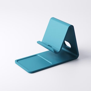 May include: A teal-colored phone stand with a curved design. The stand features a rectangular base with a raised edge and a curved support for holding a phone. A circular hole is visible on the side of the stand.