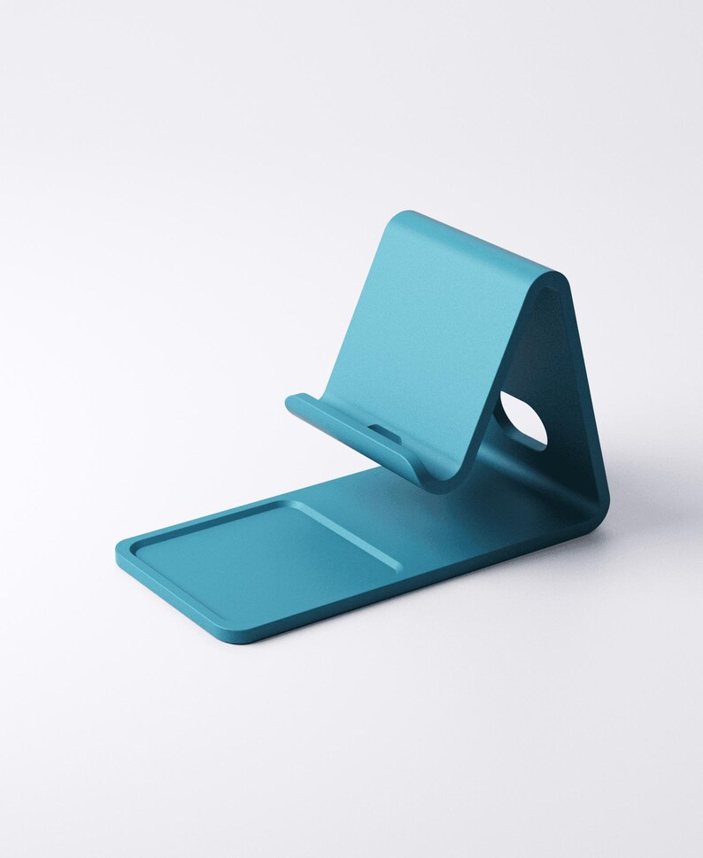 May include: A teal-colored phone stand with a curved design. The stand features a rectangular base with a raised edge and a curved support for holding a phone. A circular hole is visible on the side of the stand.