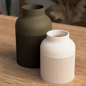 May include: Two decorative vases. One is olive green, the other is off-white. Both have a cylindrical base with vertical grooves and a smooth, rounded top. They sit on a wooden surface, showcasing a minimalist design.