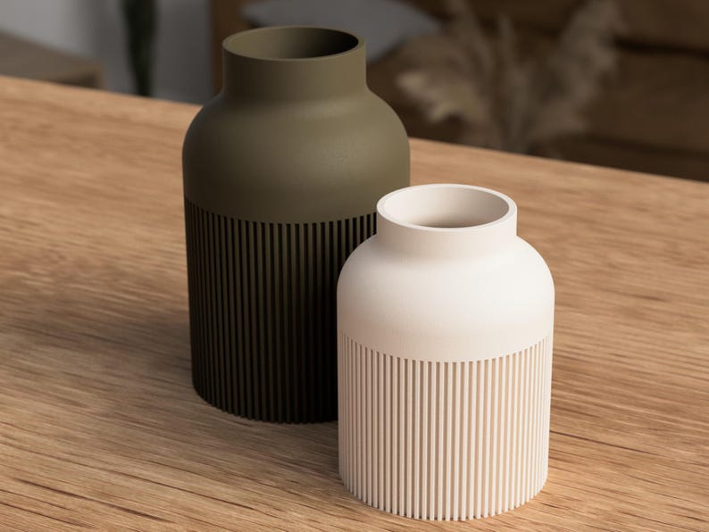 May include: Two decorative vases. One is olive green, the other is off-white. Both have a cylindrical base with vertical grooves and a smooth, rounded top. They sit on a wooden surface, showcasing a minimalist design.