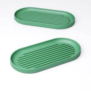 May include: Two green, oval-shaped trays. Each tray has a ribbed interior and a textured, vertical-lined border. The trays are a solid, muted green color and are set against a white background. The design is minimalist and modern.