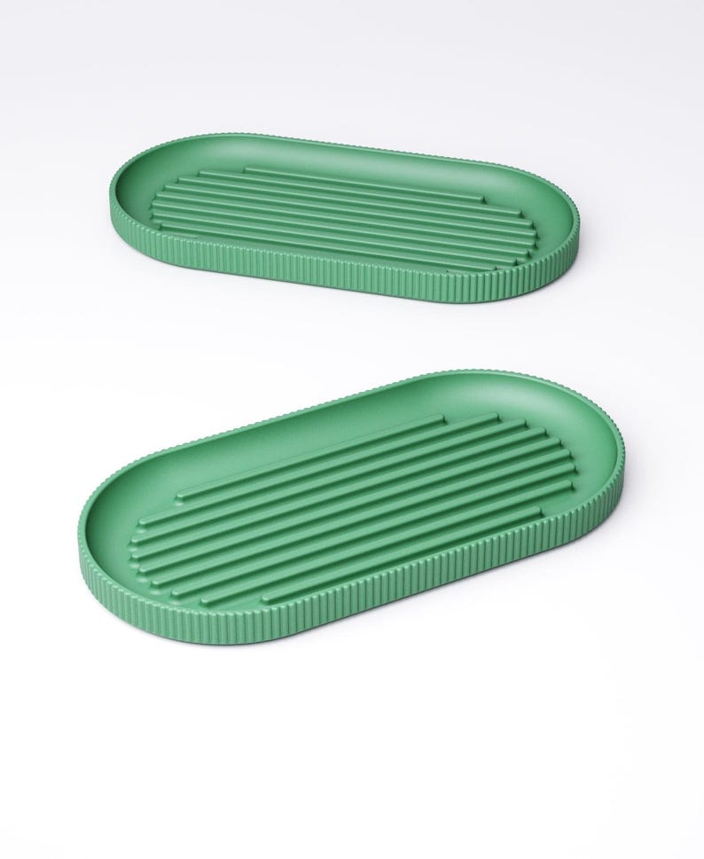 May include: Two green, oval-shaped trays. Each tray has a ribbed interior and a textured, vertical-lined border. The trays are a solid, muted green color and are set against a white background. The design is minimalist and modern.