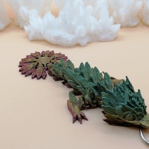May include: A detailed, multi-colored dragon keychain. The dragon features a green and brown body with a detailed head and tail. The keychain is attached to a silver metal ring. White crystal formations are in the background.
