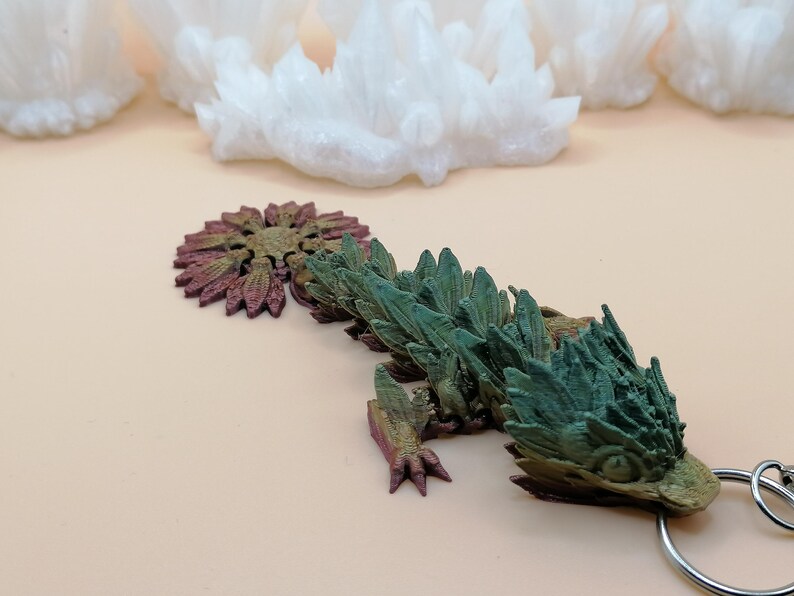 May include: A detailed, multi-colored dragon keychain. The dragon features a green and brown body with a detailed head and tail. The keychain is attached to a silver metal ring. White crystal formations are in the background.