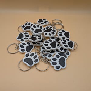 May include: A collection of white and black paw print keychains with silver key rings. The keychains are made of a smooth material and feature a detailed paw print design. The background is a solid, light tan color.