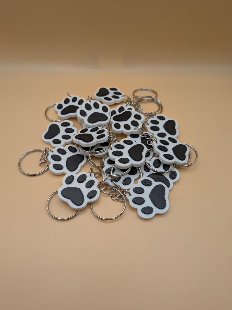 May include: A collection of white and black paw print keychains with silver key rings. The keychains are made of a smooth material and feature a detailed paw print design. The background is a solid, light tan color.