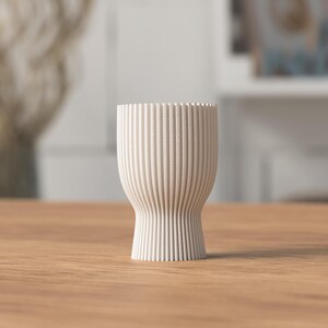 Pu&ograve; includere: Un vaso in ceramica color crema, a coste, con base svasata e parte superiore cilindrica. Il vaso &egrave; alto circa 13 cm e poggia su una superficie in legno. Le linee verticali creano un effetto testurizzato.