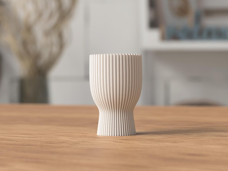 Pu&ograve; includere: Un vaso in ceramica color crema, a coste, con base svasata e parte superiore cilindrica. Il vaso &egrave; alto circa 13 cm e poggia su una superficie in legno. Le linee verticali creano un effetto testurizzato.